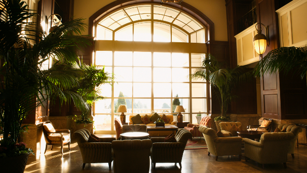 Foto einer Hotellobby; große Fensterfront, hohe Raumdecke, einige palmenartige Zimmerpflanzen.